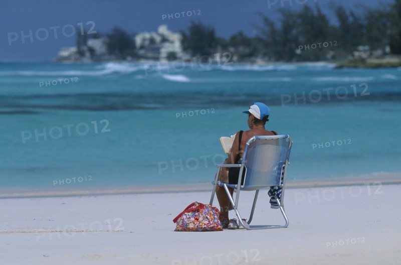 WEST INDIES, Barbados, Worthing, Woman sitting in a deck chair reading on Sandy Beach