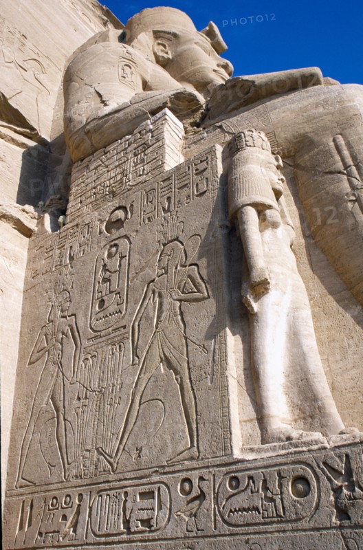 EGYPT, Nile Valley, Abu Simbel, Sun Temple of Ramses II.  Part view of one of the colossi of Ramses II flanking entrance to temple showing wall with relief carving and hieroglyphics.