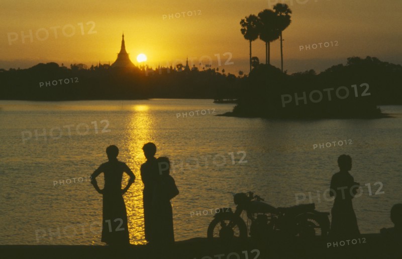 MYANMAR, Yangon, People silhouetted on shore of lake looking across towards the Shwedagon Pagoda at sunset.
