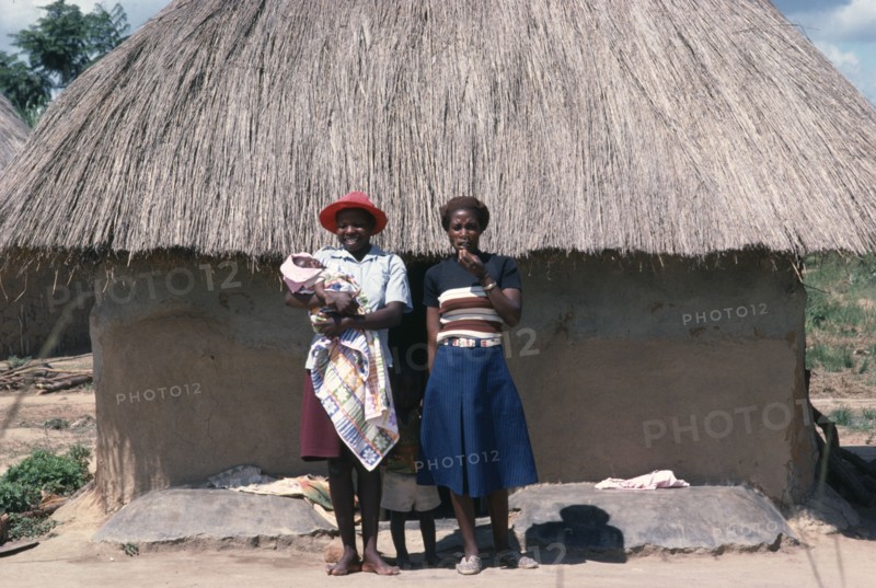 ZIMBABWE, General, Young Shona women and children standing outside thatched hut.