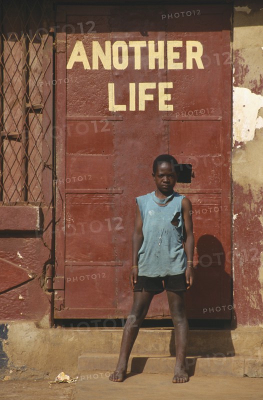 UGANDA, Kampala, Young boy standing by a red door under the words Another Life