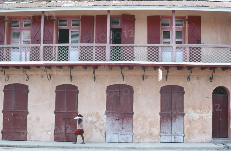 HAITI, Cap Haitien, Street scene with woman walking past building with faded pink plaster walls and red doors.