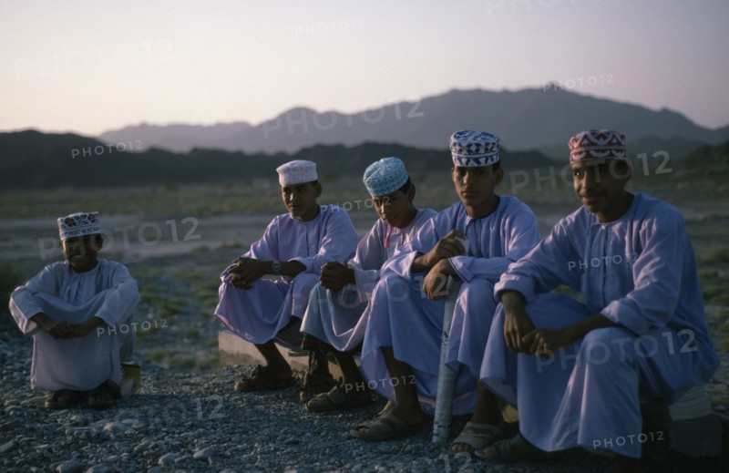 OMAN, Children, "Schoolboys wearing traditional white dishdashas and brimless, embroidered circular hats sitting outside near Bahla"