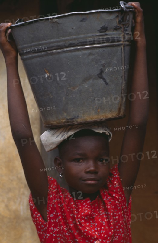 GHANA, People Carrying Goods, Portrait of child carrying large metal pail of water on her head.