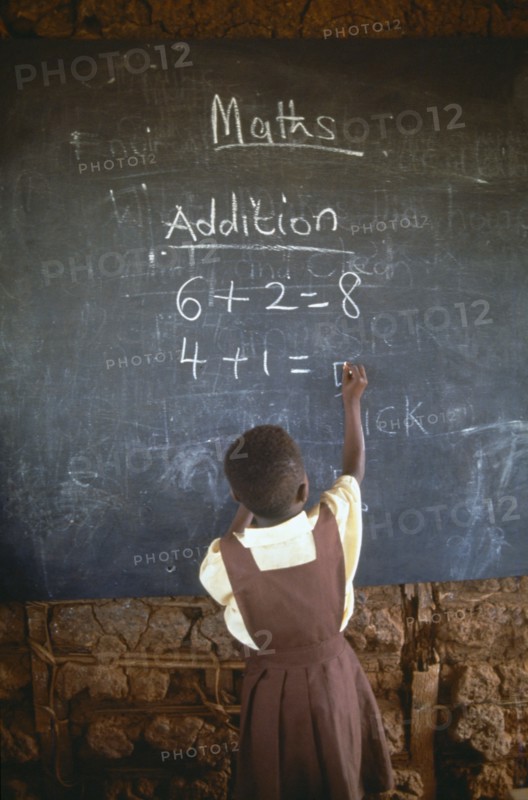 GHANA, West, Education, Child in primary school maths class writing sums on blackboard with chalk.