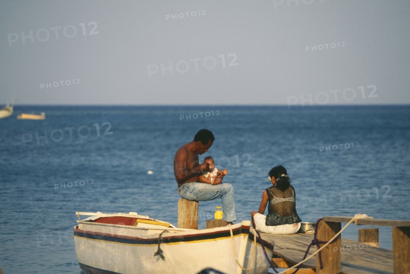 HONDURAS, Bay Islands, Roatan, Local couple with a baby sitting on wooden jetty at West End with small rowing boat tied to it.