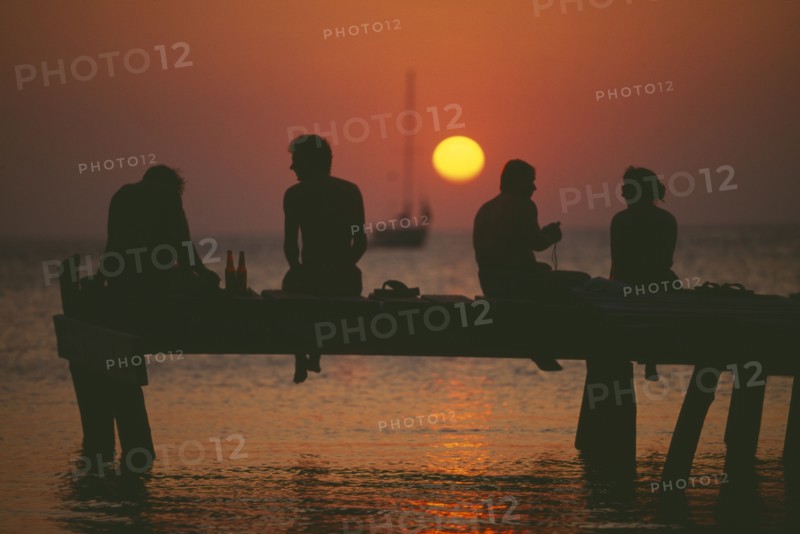 HONDURAS, Bay Islands, Roatan, Tourists sitting on jetty at West End silhouetted against warm orange sunset.