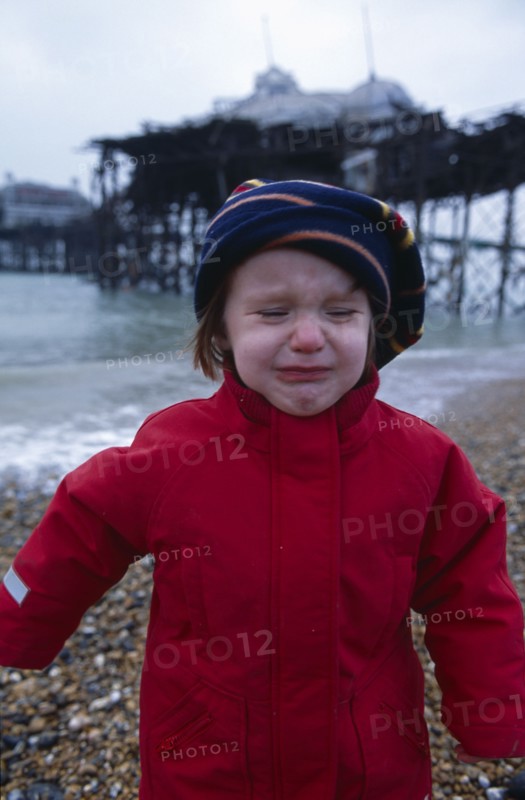 CHILDREN, Playing, Toddler dressed in red coat crying on Brighton Beach with view of the crumbling pier behind.