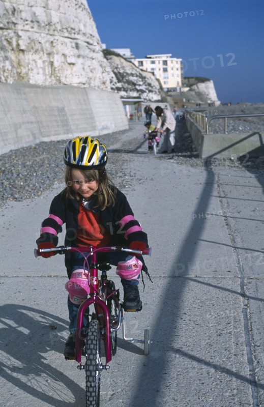 CHILDREN, Playing, Young girl on a bicycle dressed in protective cycling helmet and pads riding on a path next to the beach.