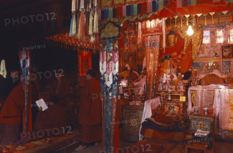 TIBET, Sakya, View though the interior of a chapel in red ambient light towards a group of monks next to a illuminated altar with figeurines displayed on it.