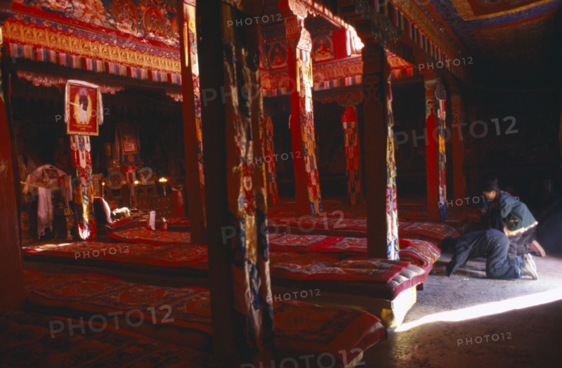 TIBET, Sakya, View through the interior of a temple in red ambient light towards the monks long cushions and Pilgrims prostrating at the entrance.