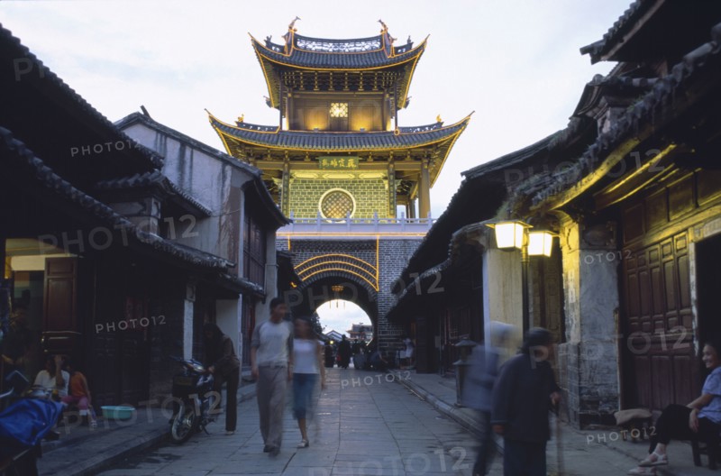 CHINA, Yunnan, Dali., Weishan. View down street at dusk towards an illuminated tower with people strolling up and down.