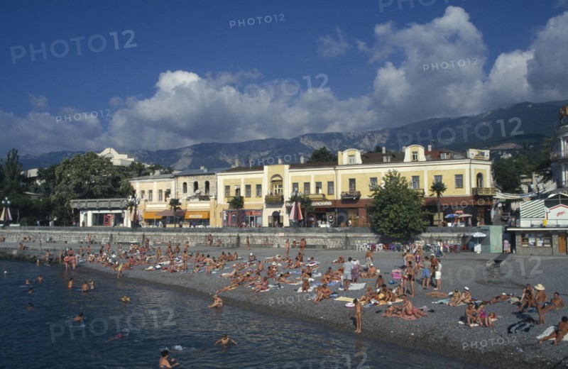 UKRAINE, Crimea, Yalta, People sunbathing on narrow beach or swimming with promenade and waterside buildings behind.