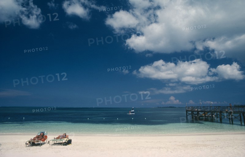 BAHAMAS, Nassau Beach, Two women sunbathing on sandy beach with windsurfer on water beyond.