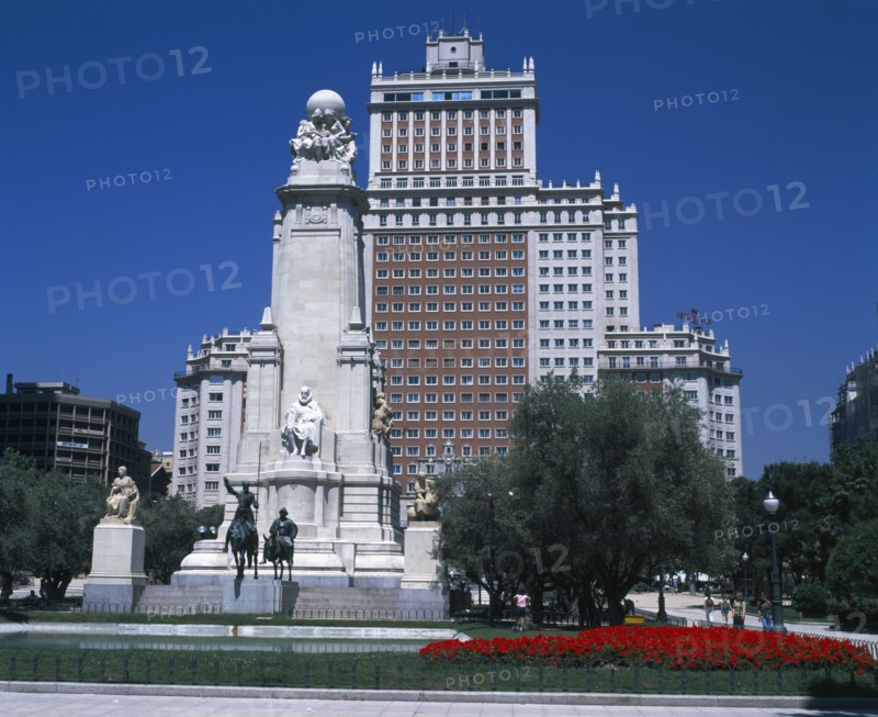 SPAIN, Madrid State, Madrid , Plaza Espana. Stone obelisk with statue of Miguel de Cervantes surrounded by trees and flower beds with tall office building behind.