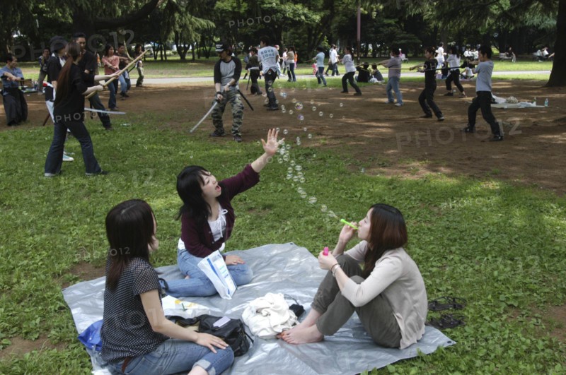 JAPAN, Honshu, Tokyo, "Harajuku. Yoyogi Park. Miki Tomiyoka, aged 23, blows bubbles for two friends while a group practices with swords in the background on a Saturday afternoon"