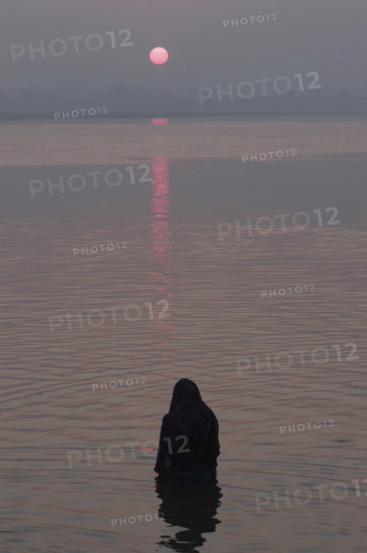 INDIA, Uttar Pradesh, Varanasi, Asi Ghat. A Hindu man baths in the Ganges River with the early morning sun over the horizon
