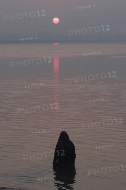 INDIA, Uttar Pradesh, Varanasi, Asi Ghat. A Hindu man baths in the Ganges River with the early morning sun over the horizon