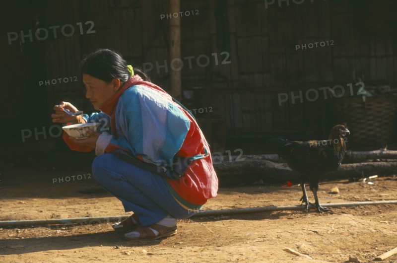 THAILAND, Chiang Rai Province, Doi Lan, Lisu woman eating whilst squatting outside her home with a chicken behind her
