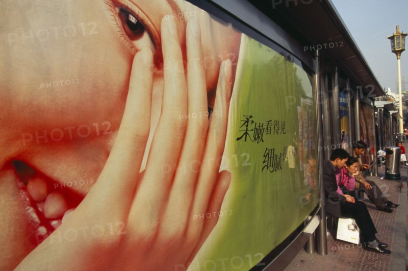 CHINA, Beijing, People waiting at bus stop south of Tiananmen Square with advertising hoarding covering exterior wall.