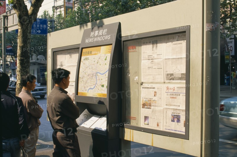 CHINA, Shanghai, Nanjing Road.  Men reading map and newspapers displayed on boards in ‘news windows’ on the street.