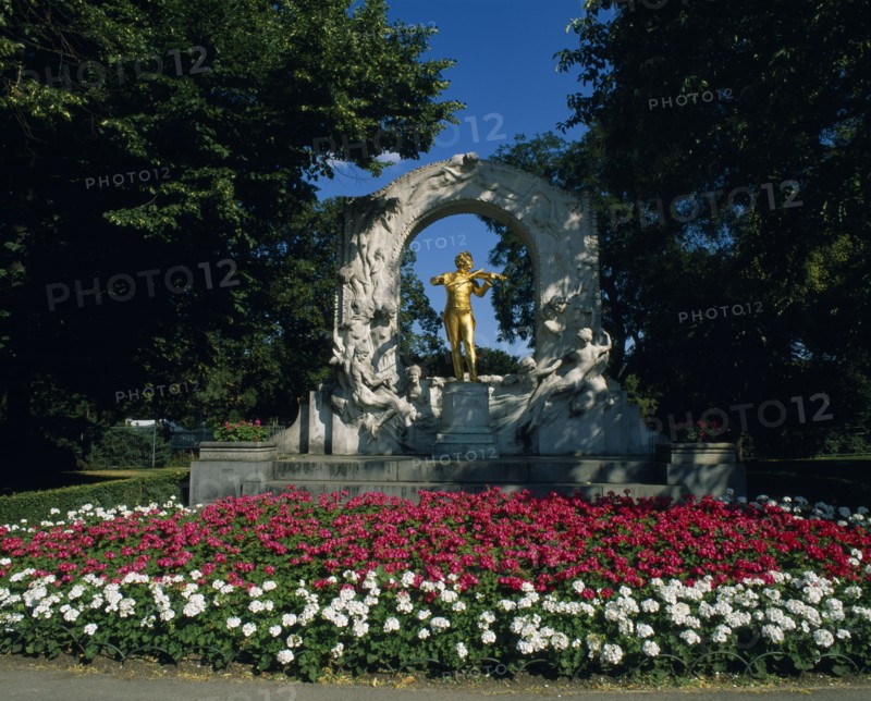 AUSTRIA, Vienna, Golden statue of Johann Strauss framed by stone arch in Stadt Park with bed of pink and white geraniums in the foreground.