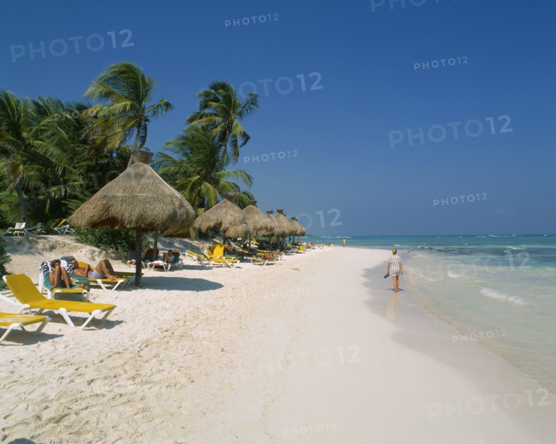 MEXICO, Quintana Roo, Tulum, Stretch of sandy beach with people on sun loungers under a line of thatched sunshades and woman walking along waters edge.