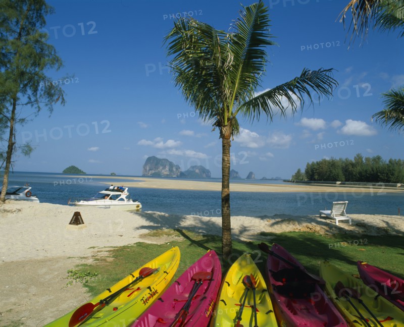 THAILAND, Trang Province, Trang Beach, Brightly coloured canoes under palm tree in foreground with view over white sand beach towards karst formations beyond.