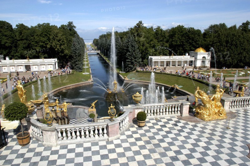 RUSSIA, Near St Petersburg, Peterhof Palace gardens. Stone balcony with chequered floor and golden statues with fountain beyond. Peterhof is also known as Petrodvorets.