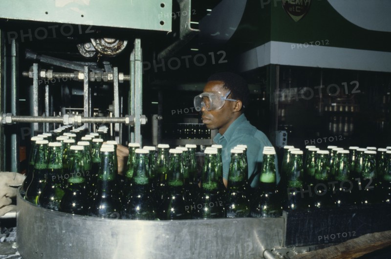 NIGERIA, Industry, Brewery with man wearing safety goggles working  behind machinery and green glass bottles.