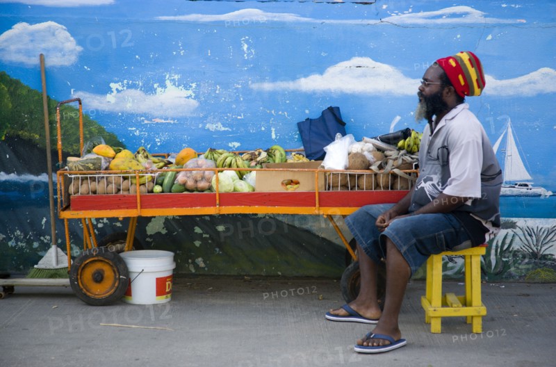 WEST INDIES, St Vincent & The Grenadines, Union Island, Fruit and vegetable stall holder in front of wall painting in Clifton