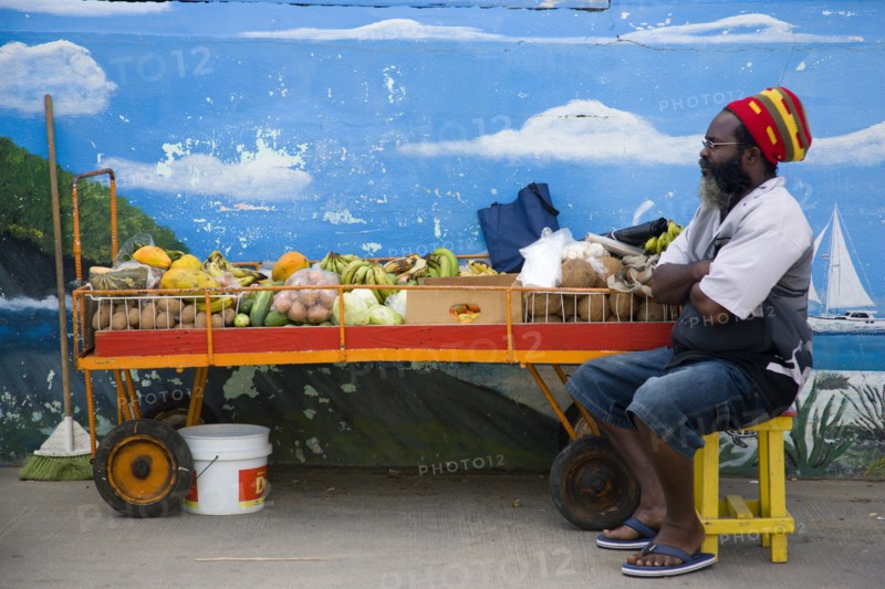 WEST INDIES, St Vincent & The Grenadines, Union Island, Fruit and vegetable stall holder in front of wall painting in Clifton