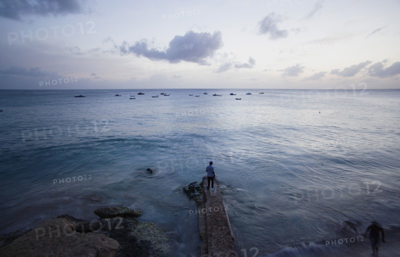 WEST INDIES, Barbados, St Lucy, Man on jetty at Half Moon Fort looking out to moored fishing boats at sunset