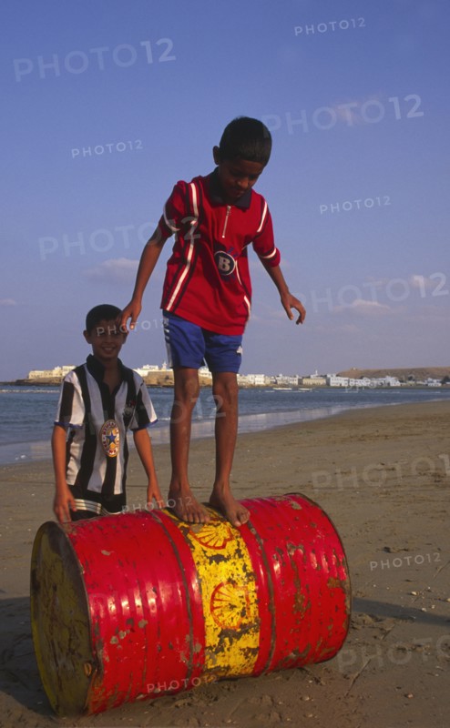 OMAN, Sur, Two young Omani boys playing on an abandoned Shell oil drum on the beach.