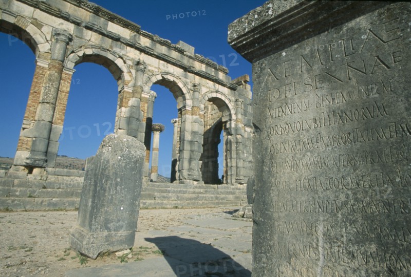 MOROCCO, Volubilis, The Basilica.  Cropped view of stone block inscribed with Roman script with arches and colonnade built into wall beyond.
