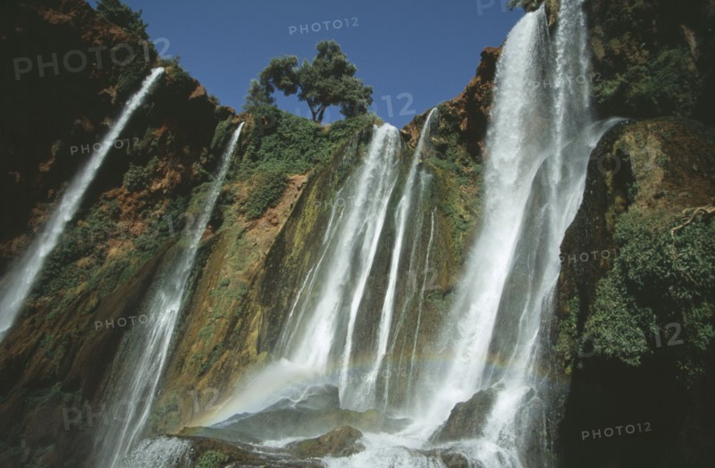 MOROCCO, Middle Atlas, Cascades d’Ouzoud, Waterfalls of the Olives.  Multiple falls cascading over rocks with sunlight through spray causing rainbow effect.
