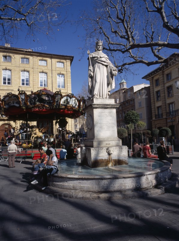 FRANCE, Aix en Provence, "Statue in centre of water fountain, people sat on edge. Carousel and buildings behind."
