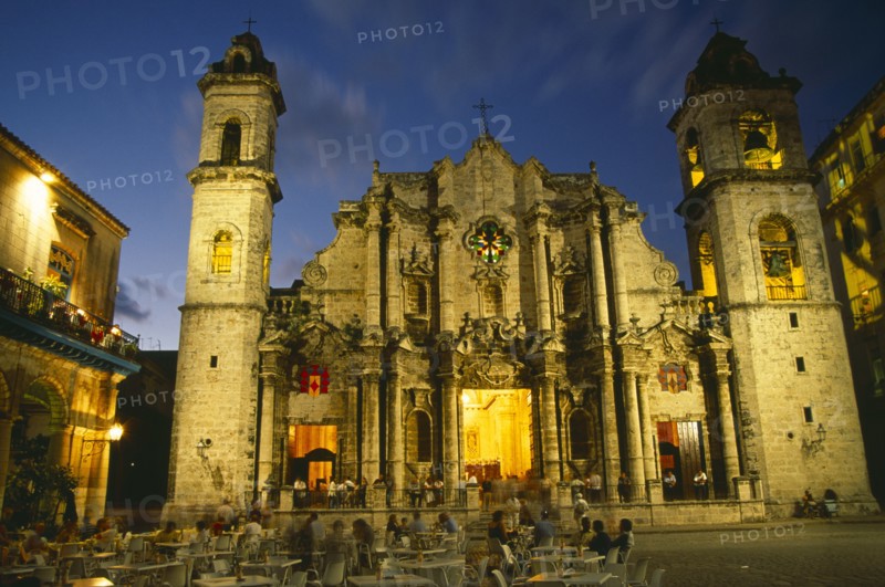 CUBA, Havana, Plaza de la Catedral.  Cathedral facade and bell tower illuminated at night with people at outside cafe tables in plaza in the foreground.