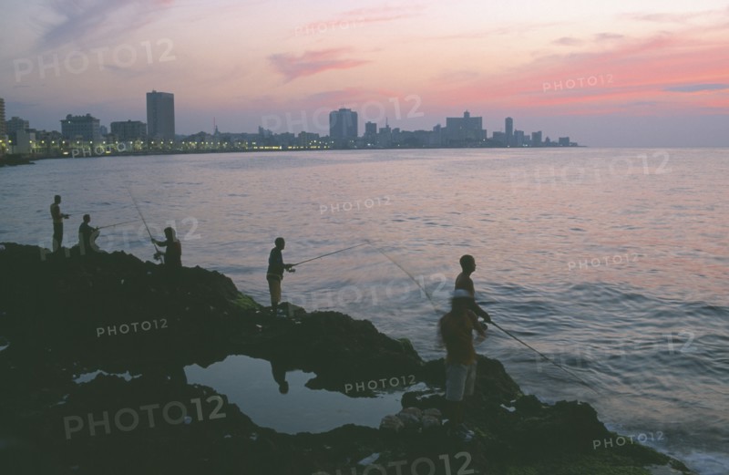 CUBA, Havana, City skyline at sunset with fishermen beside the Malecon silhouetted against pink and purple sky reflected in the sea in the foreground.