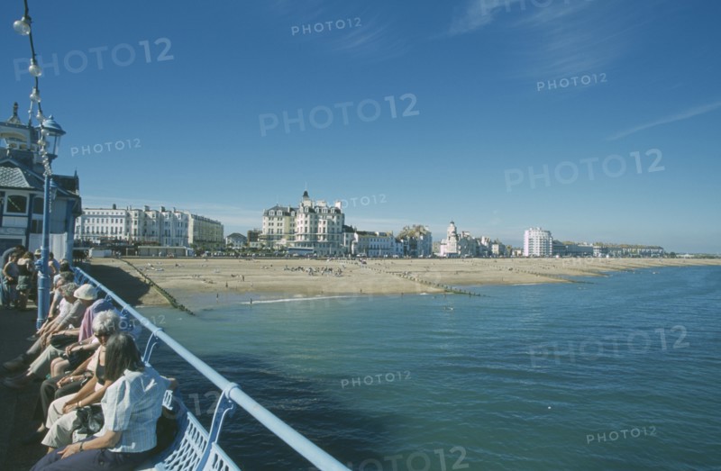 ENGLAND, East Sussex, Eastbourne, View from pier with people sat on seats towards beachfront.