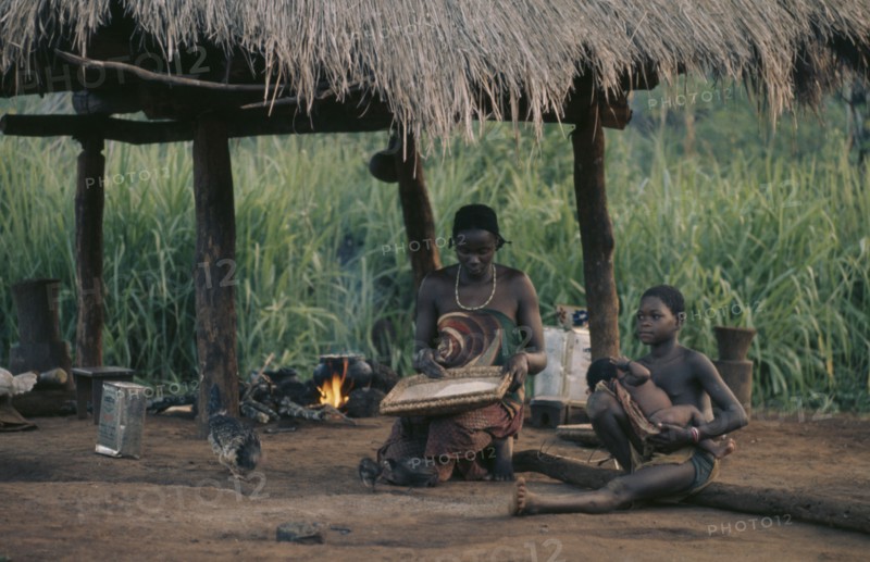 CONGO, Tribal People, Woman and children sitting beneath thatched shelter in village with pot on open fire behind.