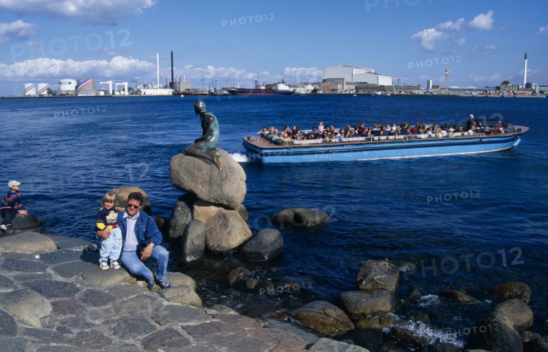 DENMARK, Zealand, Copenhagen, Family posing for photograph beside the Little Mermaid statue with passing tourist boat behind and city skyline beyond.