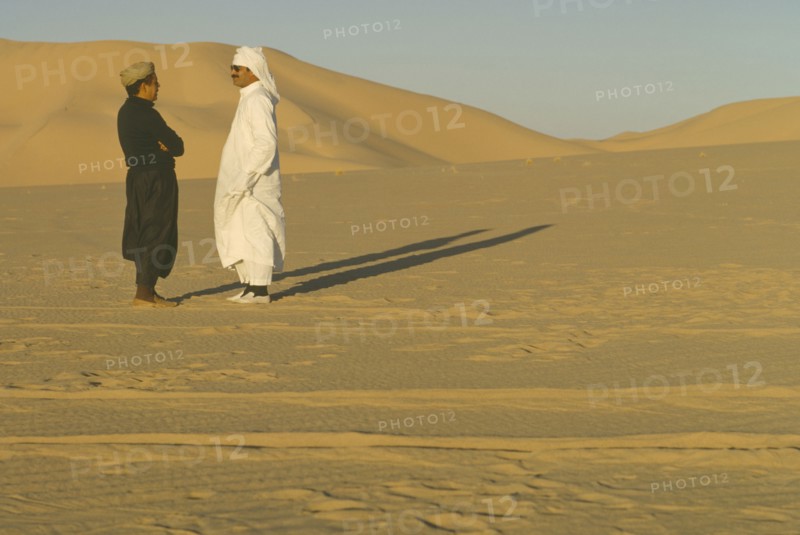 ALGERIA, Desert, Two men standing in the desert in conversation casting strong shadows across the sand.