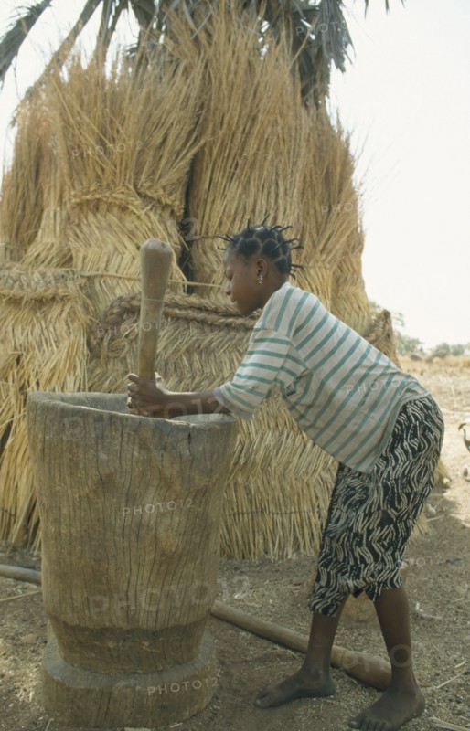 BURKINA  FASO, Bisaland, Sigue Voisin, Near Garango. Young girl pounding grain with straw roofs and granaries behind