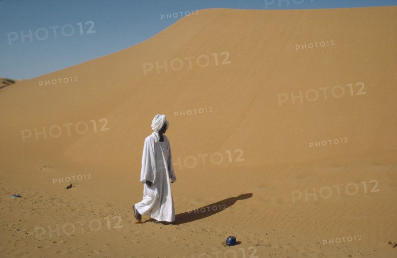 SUDAN, Kordofan Province, El Humra Village, Man in white robe and turban passing huge sand dunes encroaching on village and agricultural land and cutting off road to El Caeid.