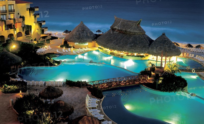 MEXICO, Jalisco ,  Puerto Vallarta, "A swimming pool surrounded by sun loungers, wooden thatched huts and bridges lit up at night. Hotel right next to beach."