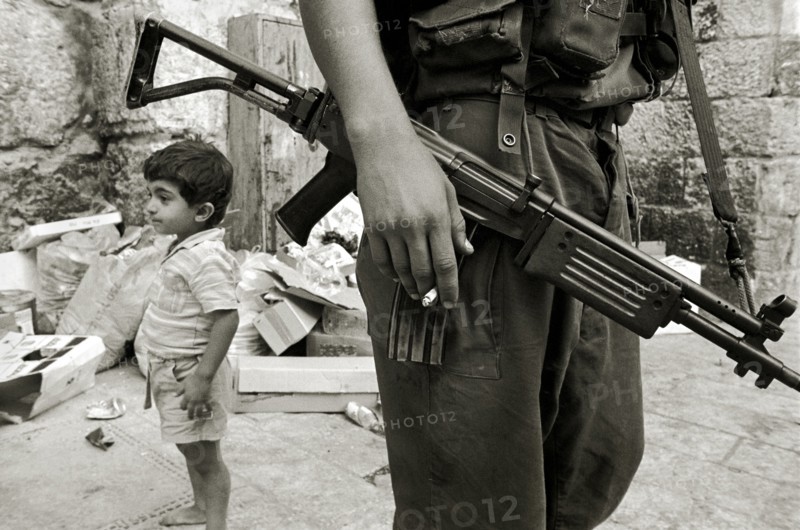 ISRAEL, Jerusalem, "Israeli soldier with rifle and cigarette relaxes in the Arab Quarter, small boy standing behind."