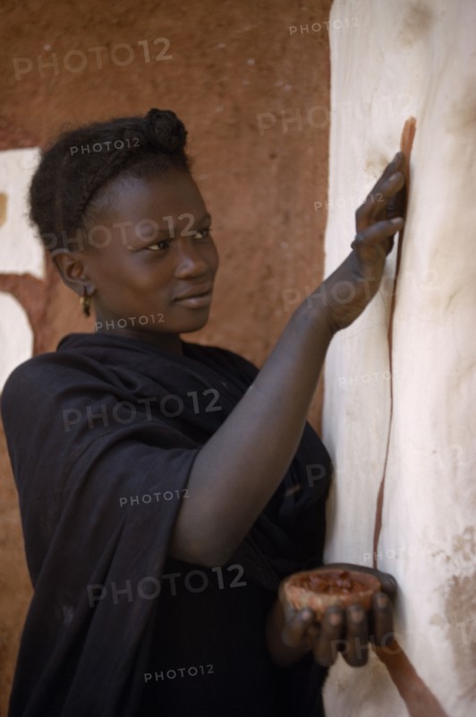 MAURITANIA, Oualata, Young woman decorating exterior wall of mud building with ochre.  It is traditional for women to both design and apply such decorations.
