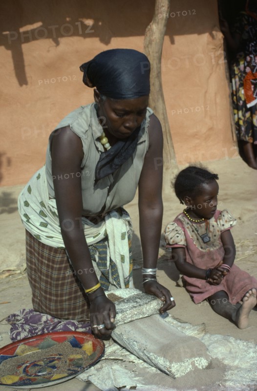KENYA, People, Women, Burji woman grinding grain on large flat stone to produce flour with child sitting beside her.