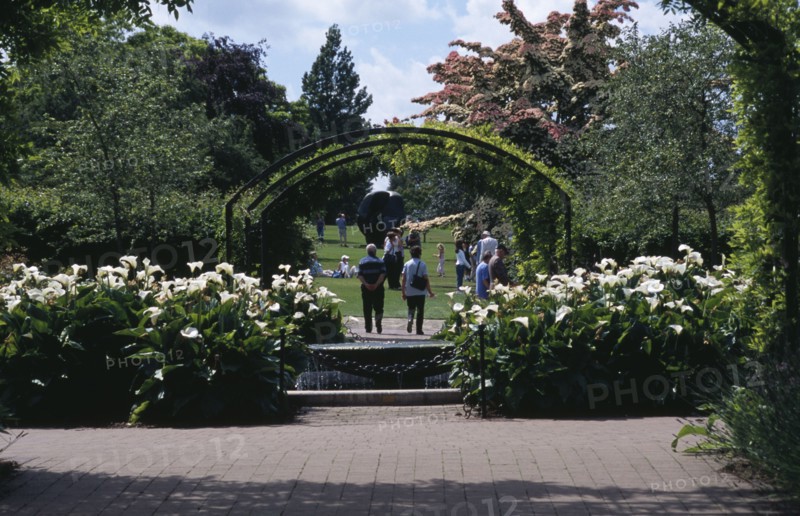 ENGLAND, Surrey, Woking, Wisley Royal Horticultural Society Garden. Visitors walking along pathway through arch towards Henry Moore sculpture.